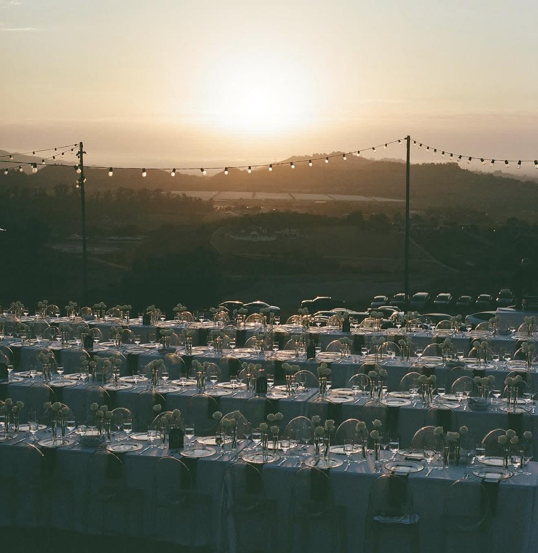 Long banquet tables set with white roses on a hillside at sunset