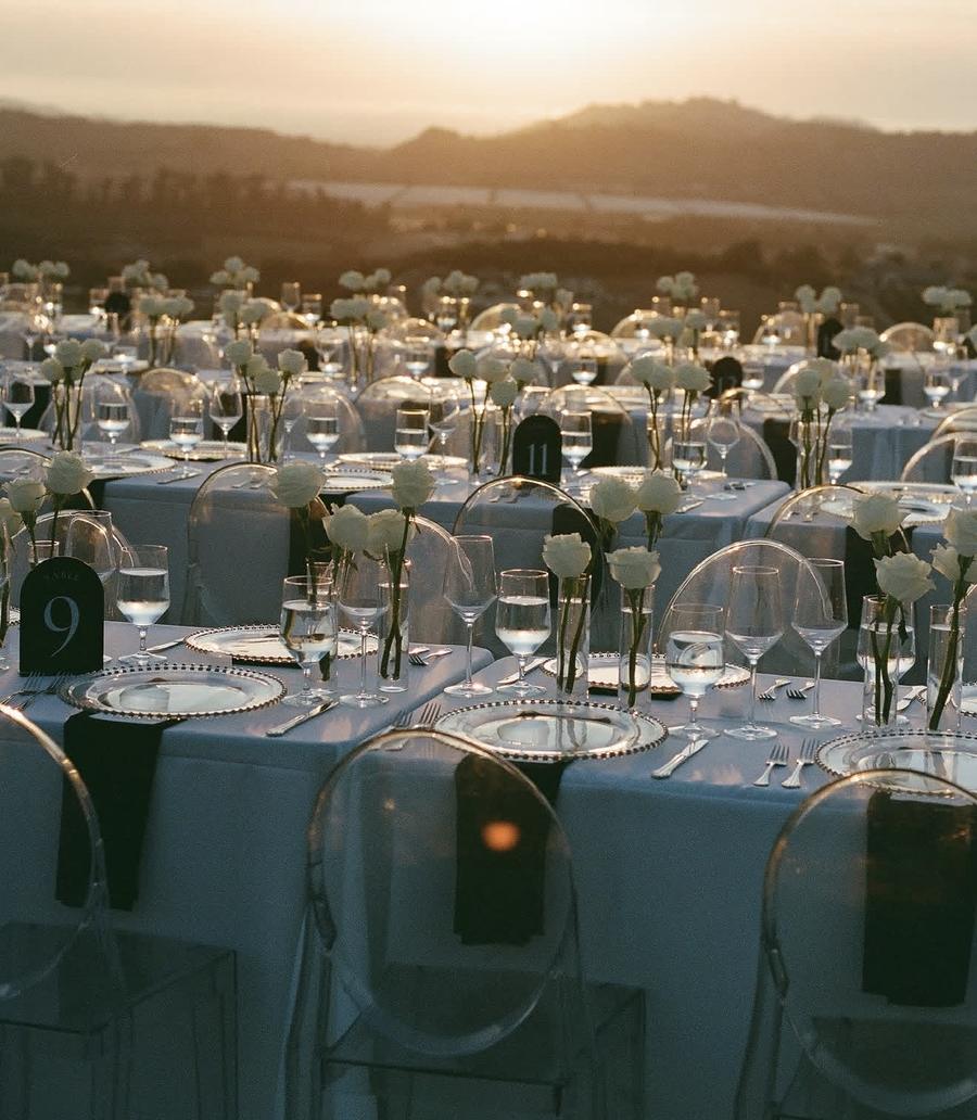 Bud vases of white roses on long tables with numbered table cards at sunset