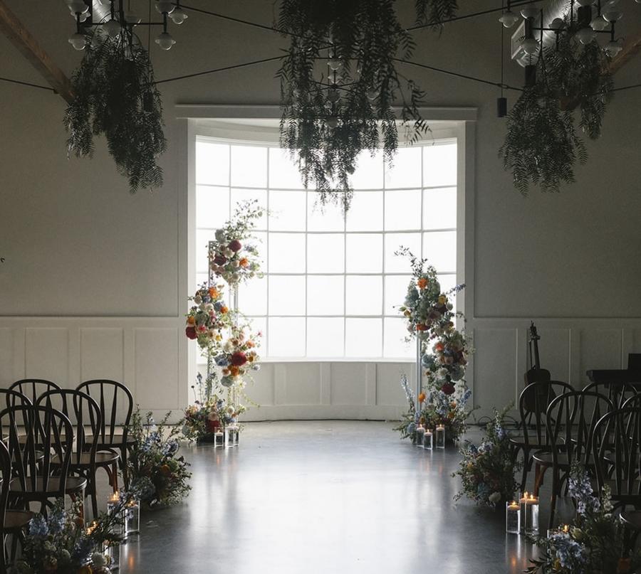 Aisle lined with wildflower arrangements and suspended greenery before a tall window